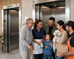 Hall térreo de condomínio residencial moderno com dois elevadores de acesso compartilhado. Grupo diverso de pessoas aguarda o elevador social de forma respeitosa e inclusiva: idoso com bengala, trabalhadora doméstica, família com criança, casal com cachorro de pequeno porte no colo representando família multiespécie, e entregador com caixa, ilustrando o fim da distinção discriminatória entre elevador social e de serviço conforme Projeto de Lei 4.710/2024 e a convivência sem discriminação.