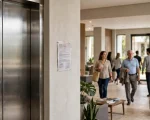 Elevador em primeiro plano, aviso institucional discreto com texto ilegível ao lado, e o hall com moradores ao fundo transmitindo convivência e responsabilidade social
